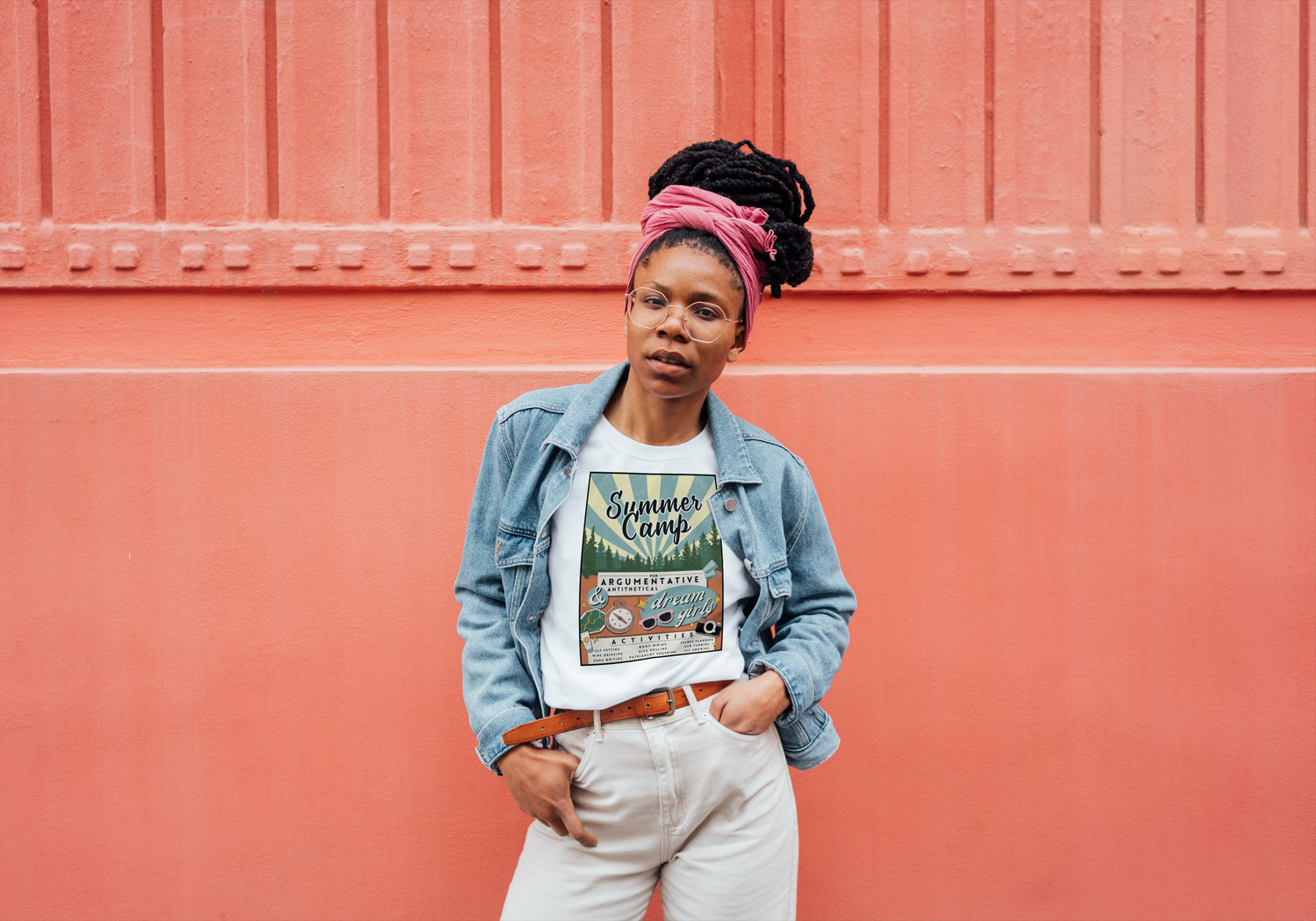 A black women stands with her hands in her pockets, looking past the camera. There is a reddish industrial wall behind her. She is wearing tan pants and a Summer Camp (Taylor's Version) shirt with a jean jacket.