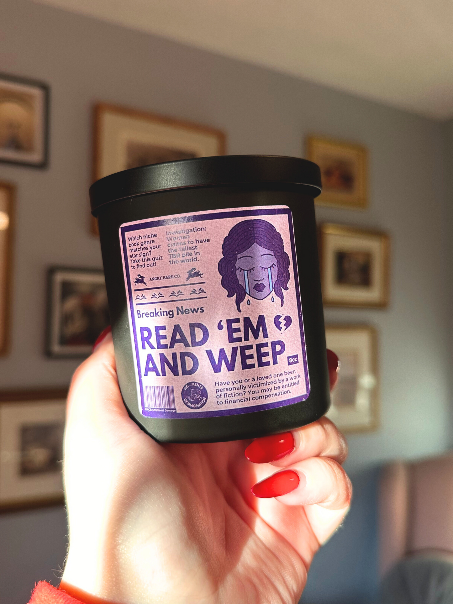 A hand holds an 8oz soy candle in a matte black jar with a purple monochrome label that resembles a magazine cover. It says, "Read 'Em And Weep."